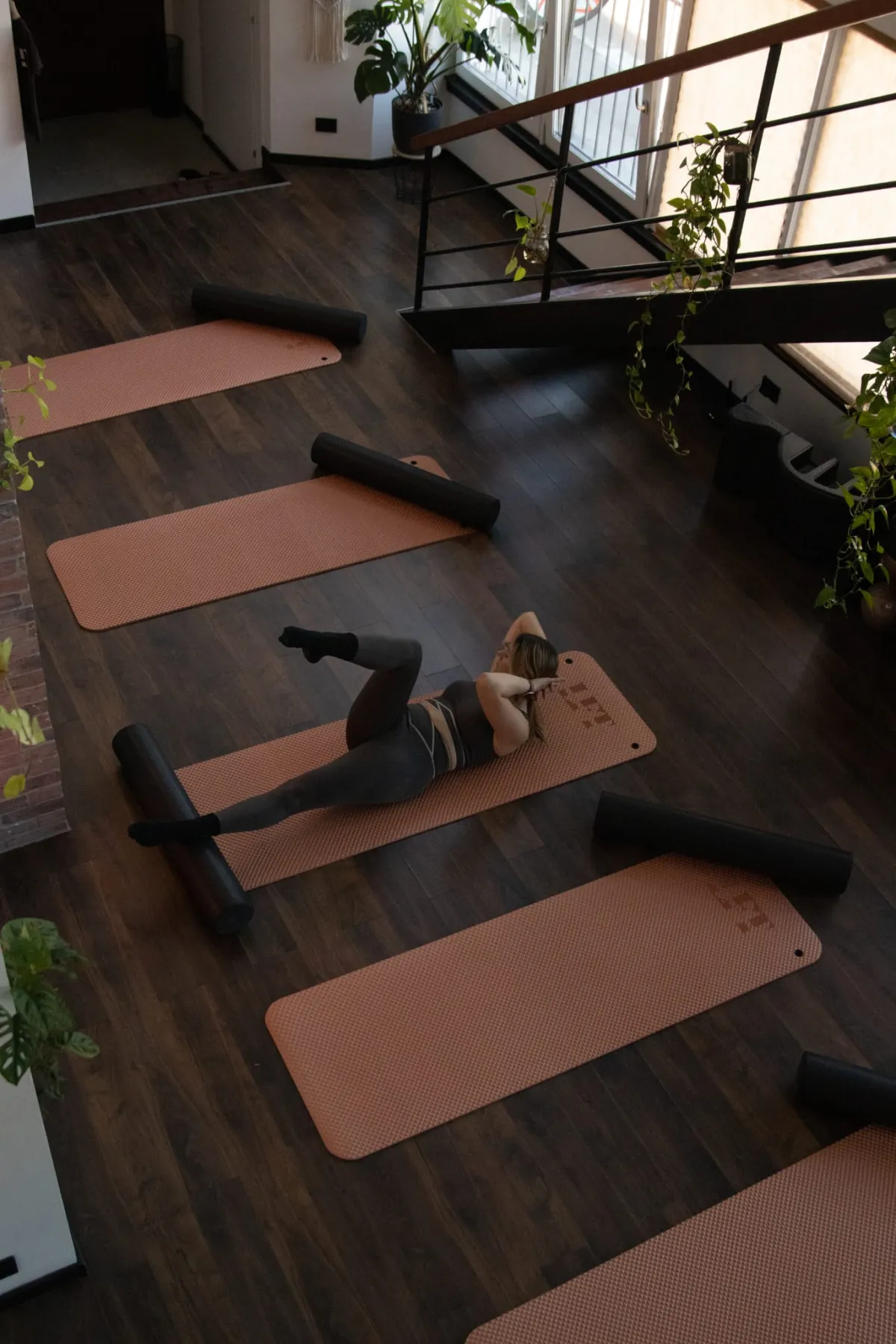 Lezione di Pilates Matwork vista dall’alto: tappetini terracotta con rullo allineati sul pavimento in legno dello studio, un’allieva a terra a metà esercizio, piante e scala sullo sfondo.